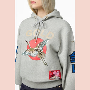 Goldbergh Tobu chest detail showing sequined crane, GOLDBERGH patch, and drawstring on heather gray hoodie
